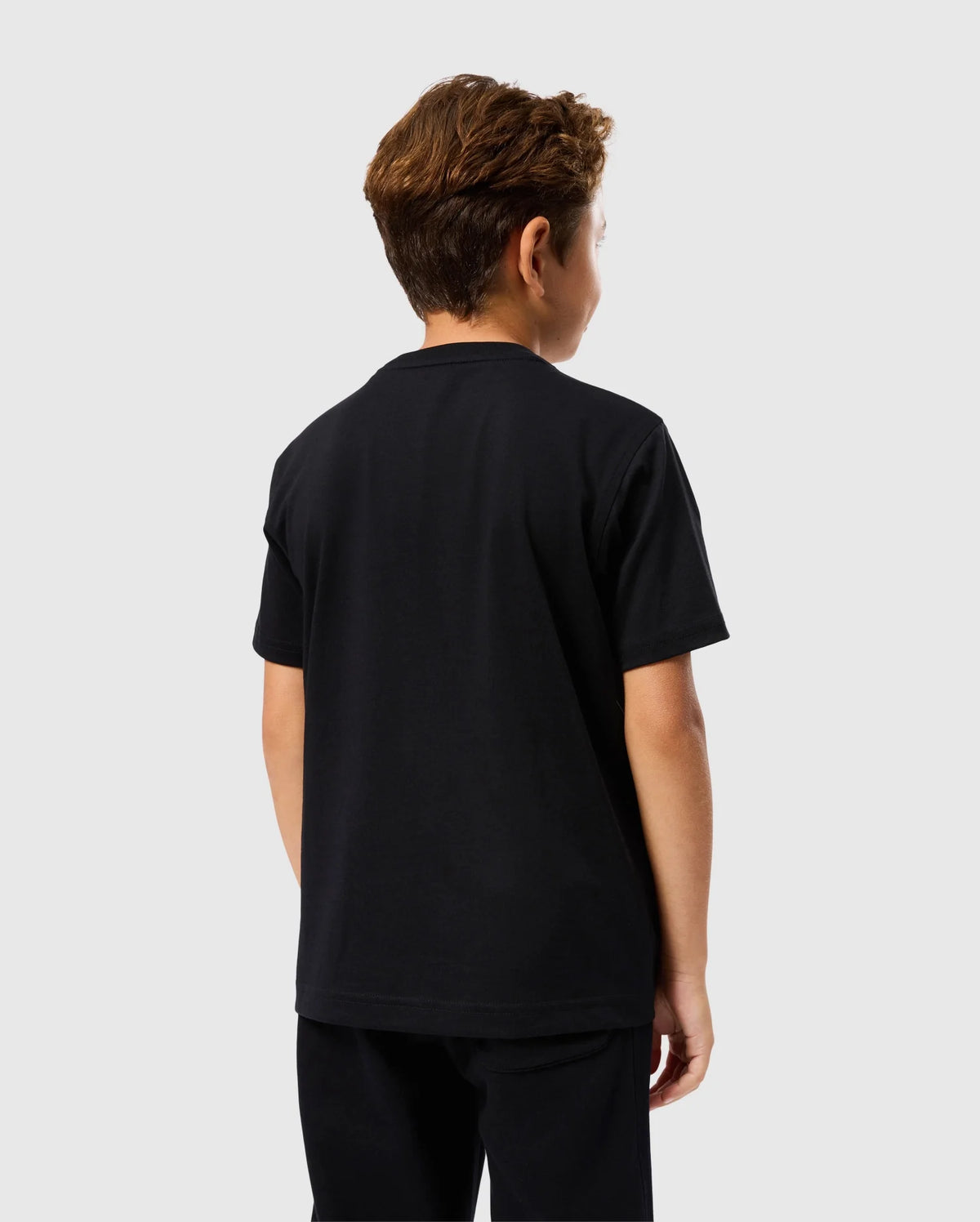 A young boy with short brown hair is standing with his back to the camera. He is wearing a black Psycho Bunny KIDS MALTON GRAPHIC TEE - B0U795D200, made of luxurious Pima cotton, along with black pants, all set against a plain white background.