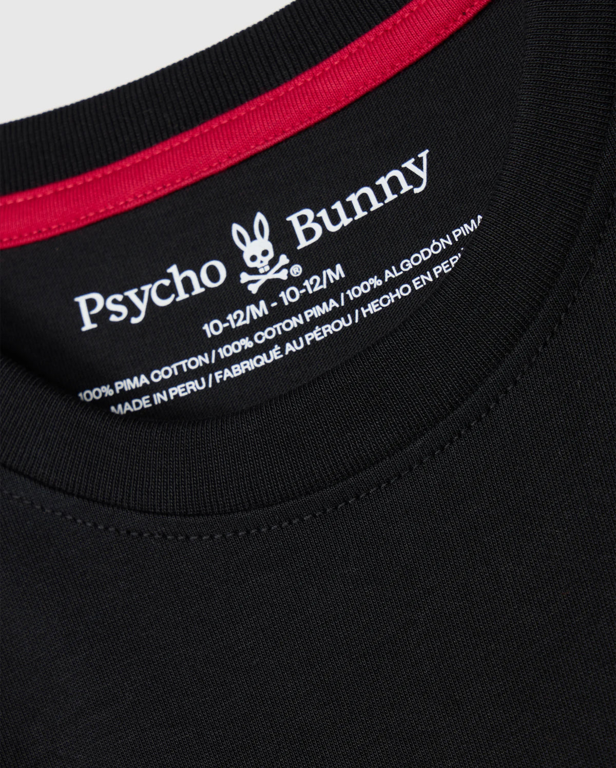 Close-up of a black KIDS MALTON GRAPHIC TEE - B0U795D200 collar with red trim. The label inside the collar displays the Psycho Bunny brand name and a bunny logo, along with size details "10-12/M" and material composition "100% Pima cotton," showcasing Peruvian craftsmanship in both English and Spanish.