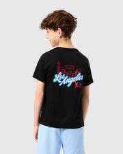 A child with curly hair, seen from behind, wears the KIDS LOS ANGELES CITY TEE - B0U861A2PC by Psycho Bunny. The soft black Peruvian Pima cotton tee showcases a vibrant graphic design of buildings and the text "Los Angeles" alongside "Psycho Bunny." Light blue shorts complete the look against a gray background.