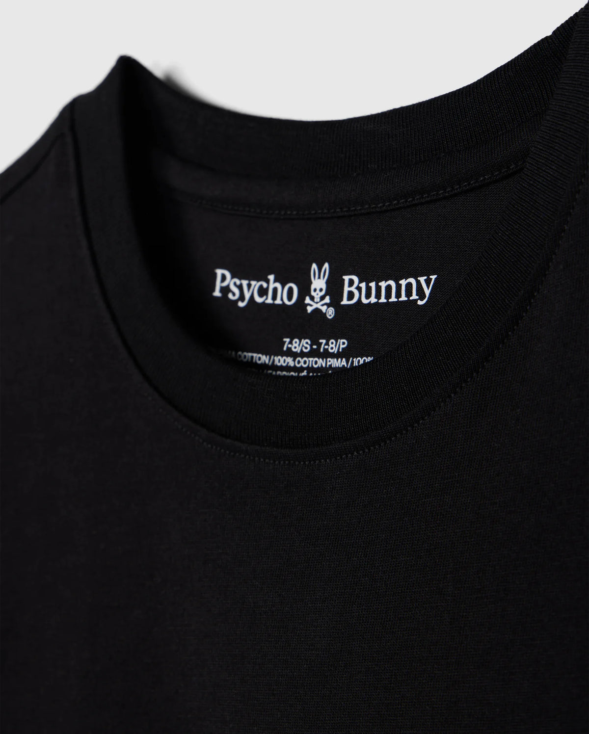 Close-up of a black t-shirt, showcasing the brand name "Psycho Bunny" and a small bunny skull logo inside the collar. Details include shirt size and material info, made from 100% Peruvian Pima cotton. The soft and smooth fabric highlights its premium quality. This particular piece is identified as the "KIDS LOS ANGELES CITY TEE - B0U861A2PC".