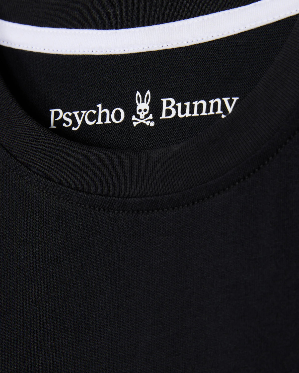Close-up of the KIDS ZACK GRAPHIC TEE (B0U897D200) from Psycho Bunny, featuring a black Pima cotton shirt collar with the brand's iconic bunny head and crossbones logo, accented by white stitching along the neckline.