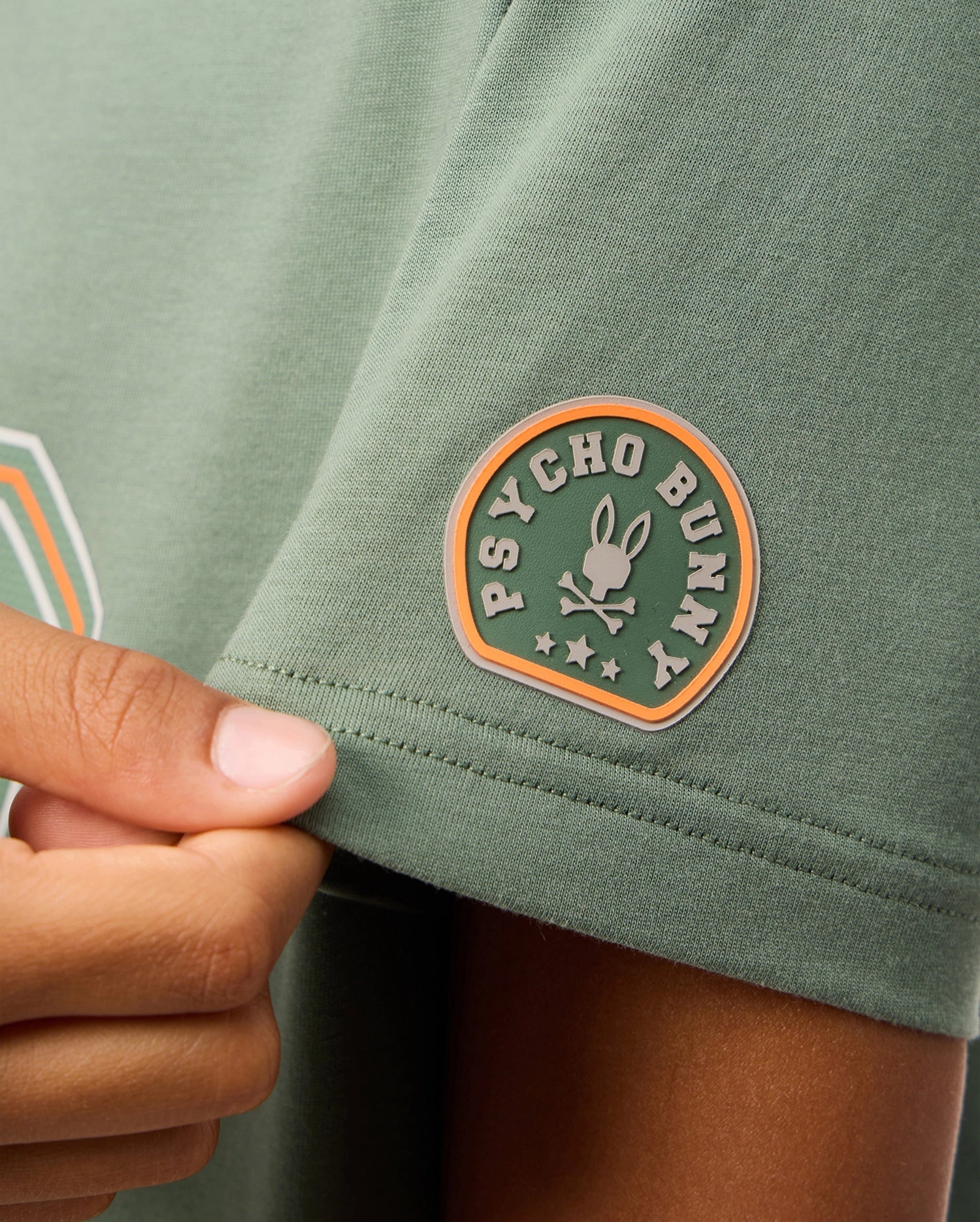 Close-up of someone holding the sleeve of a green KIDS ZACK GRAPHIC TEE by Psycho Bunny. The sleeve features a circular patch displaying "Psycho Bunny" and a bunny skull logo encircled by stars, made from high-quality Pima cotton for exceptional comfort.