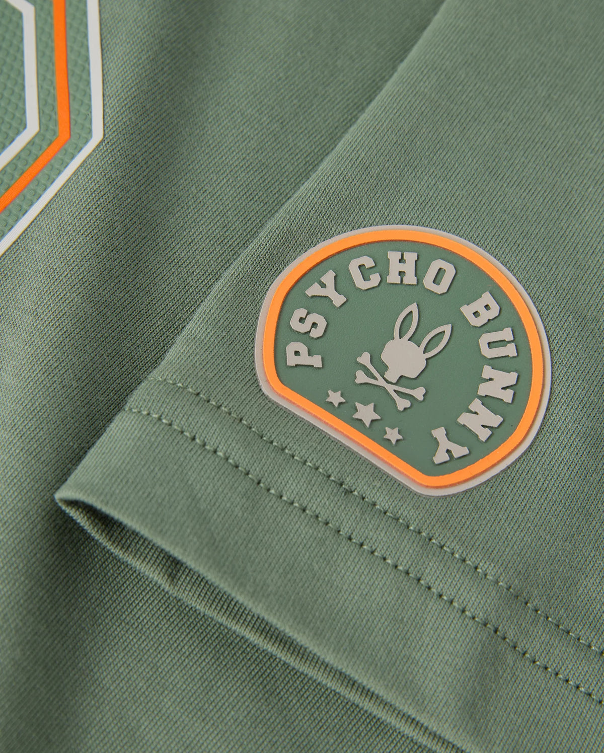 Close-up of the green Pima cotton fabric of the KIDS ZACK GRAPHIC TEE - B0U897D200, highlighting a logo that features a bunny silhouette, two bones, and stars. The design is encircled with "Psycho Bunny" in white and includes vibrant orange and white accents, making it ideal for this distinctive graphic tee from Psycho Bunny.