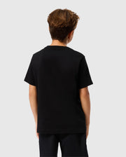 A young person with short brown hair is wearing the KIDS TACOMA GRAPHIC TEE - B0U909D200 by Psycho Bunny and black pants featuring subtle embroidered details, as they face away from the camera against a light gray background.