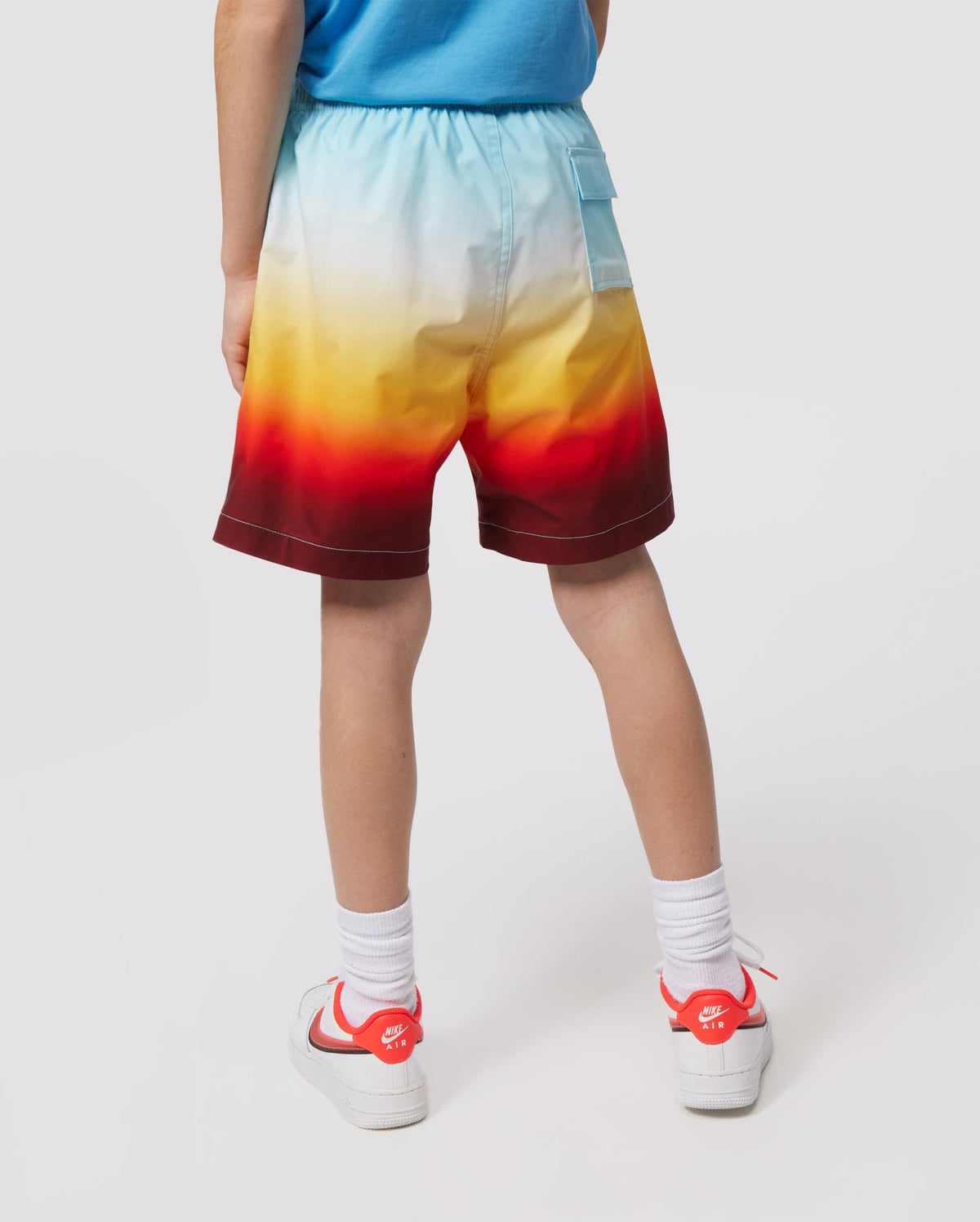 A child from the waist down wearing vibrant print Psycho Bunny kids' swim trunks transitioning from blue to red, paired with white socks and red sneakers, standing against a white background.