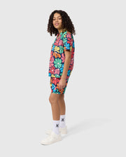 A person with curly hair wearing a colorful, floral-pattern shirt and matching KIDS MENTZ SWIM TRUNK by Psycho Bunny stands sideways. They are smiling and looking at the camera. They are also wearing white socks with a small symbol on them and white sneakers. The background is plain.