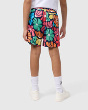 A person wearing a white t-shirt and the KIDS MENTZ SWIM TRUNK by Psycho Bunny, featuring a colorful, floral pattern, stands facing away from the camera. They are also wearing white socks and white sneakers. The background is plain gray.
