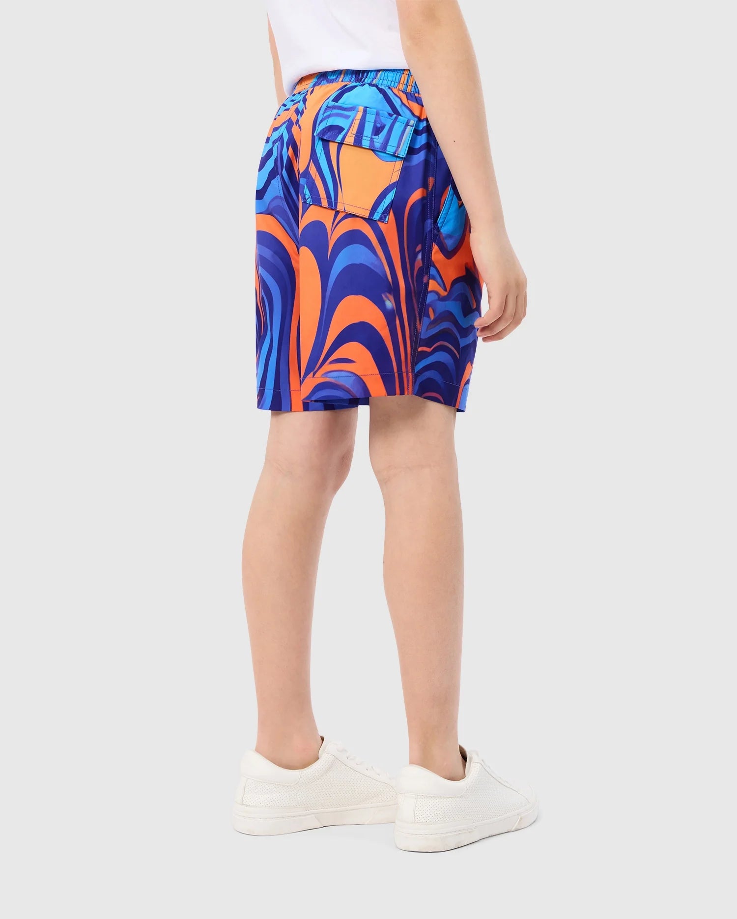 A person is standing, facing away, wearing a white shirt and KIDS NEVADA ALL OVER PRINT SWIM TRUNK - B0W526C200 from Psycho Bunny, made from quick-dry fabric with colorful blue and orange patterns, paired with white sneakers. The background is plain light gray.