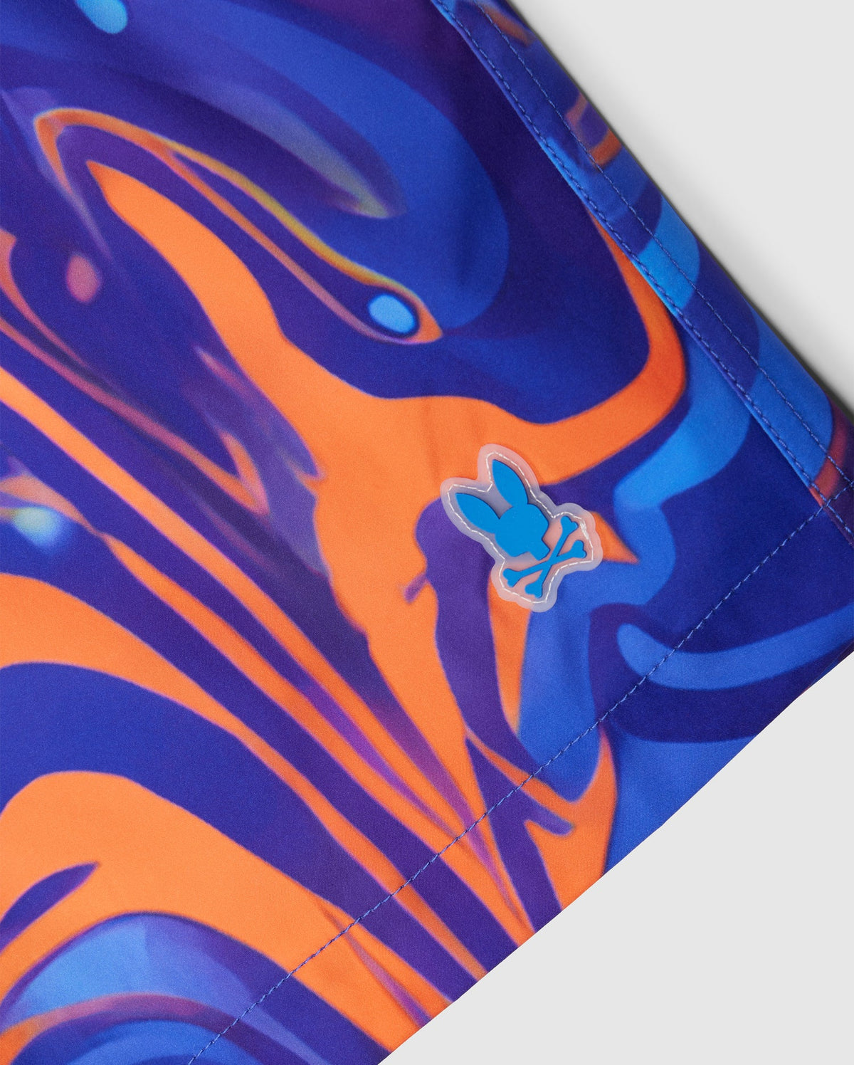 Close-up of the KIDS NEVADA ALL OVER PRINT SWIM TRUNK - B0W526C200 by Psycho Bunny, showcasing a vibrant abstract swirl pattern in blue, purple, and orange hues. Made from quick-dry fabric, these trunks feature a small embroidered logo near the edge depicting a stylized light blue rabbit with crossed bones beneath it.