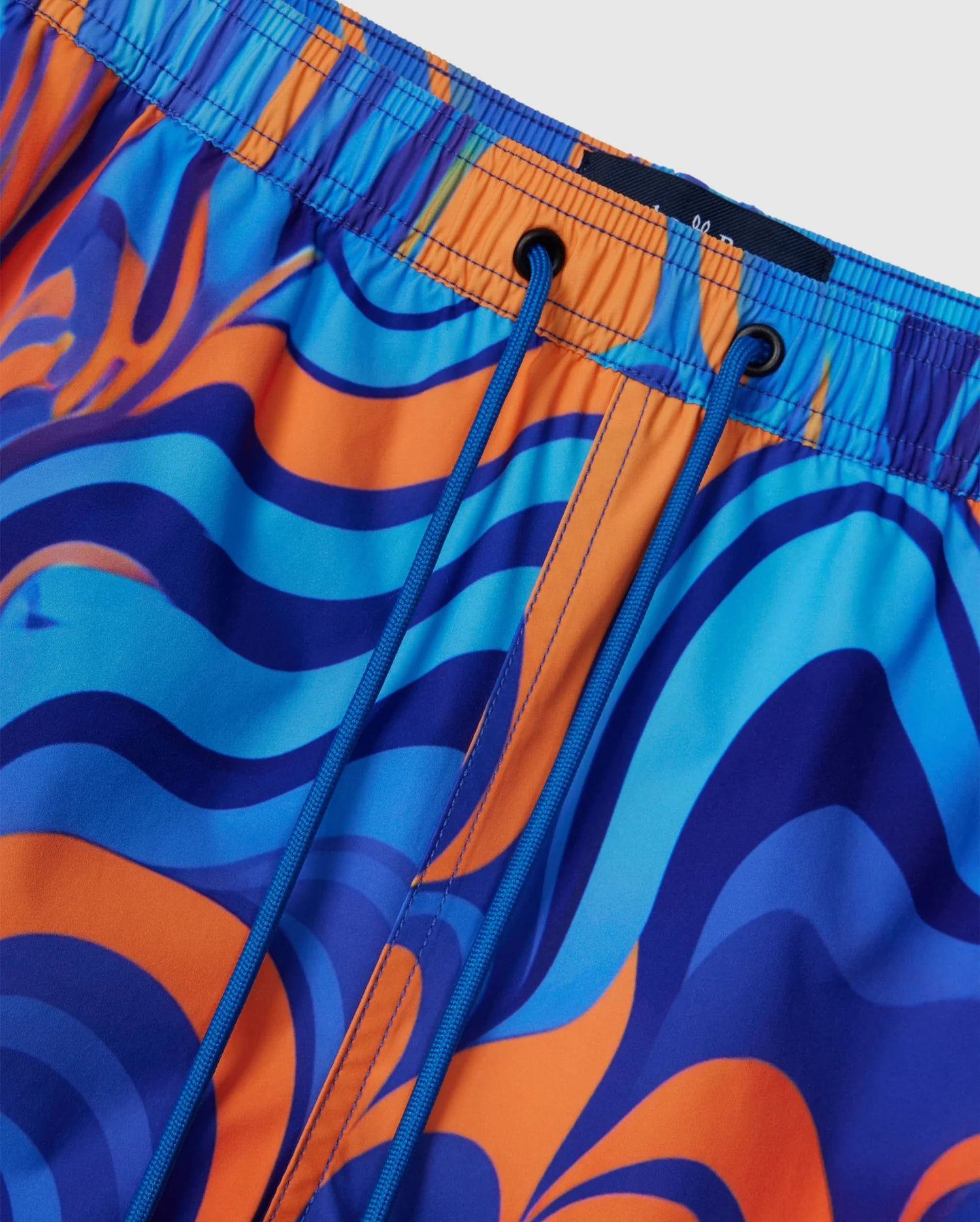 Close-up of the Psycho Bunny KIDS NEVADA ALL OVER PRINT SWIM TRUNK (B0W526C200), featuring an abstract, wavy pattern in shades of blue, orange, and purple. These quick-dry fabric shorts come with an elastic waistband and blue drawstrings.