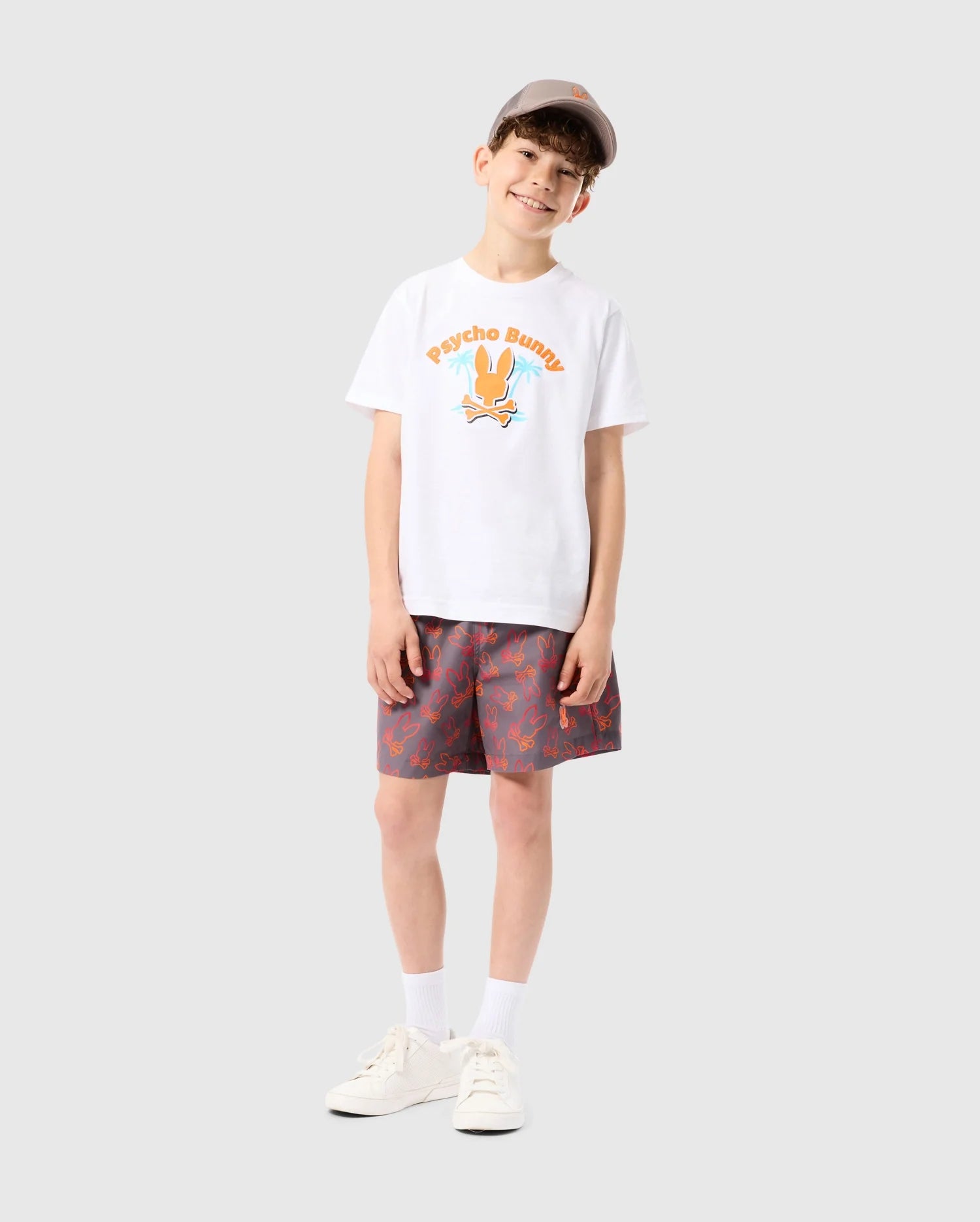 A smiling young boy stands against a plain background, wearing a white t-shirt with colorful graphics, KIDS SHELDON ALL OVER PRINT SWIM TRUNK - B0W588C200 from Psycho Bunny, white socks, and sneakers. He is also wearing a beige cap, and his hands are relaxed at his sides.