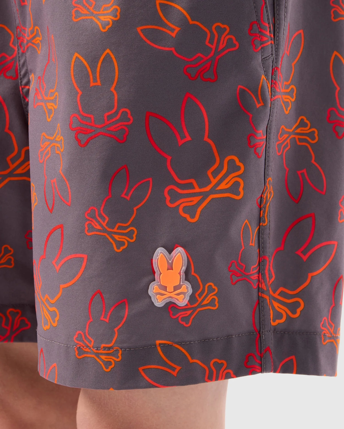 Close-up of Psycho Bunny KIDS SHELDON ALL OVER PRINT SWIM TRUNK - B0W588C200 featuring a repetitive bunny pattern of skulls in red and orange outlines on dark-colored, quick-dry fabric. The shorts have a small, clear bunny skull emblem near the hem, with a portion of a child's leg visible at the bottom.
