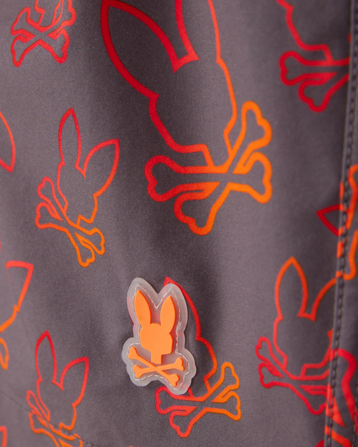 Close-up of Psycho Bunny KIDS SHELDON ALL OVER PRINT SWIM TRUNK - B0W588C200 featuring a bunny pattern with skulls and crossbones in red, orange, and yellow. There's a small rubberized patch of the same bunny skull and crossbones design in orange attached to the quick-dry fabric. The background is a muted gray color.