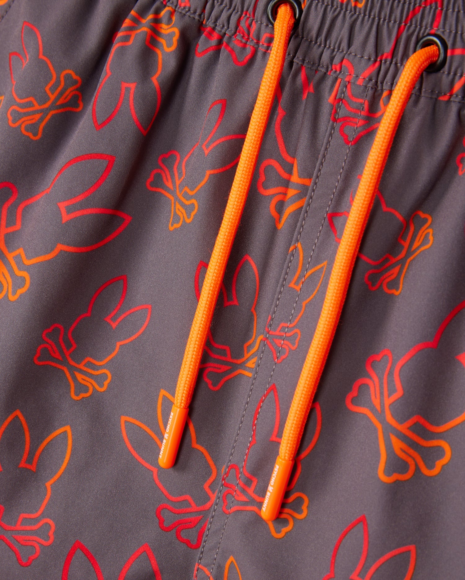 Close-up of gray quick-dry fabric with an elastic waistband and bright orange drawstrings. The Psycho Bunny KIDS SHELDON ALL OVER PRINT SWIM TRUNK - B0W588C200 is patterned with red and orange bunny skull and crossbones designs. The drawstrings have matching orange aglets.