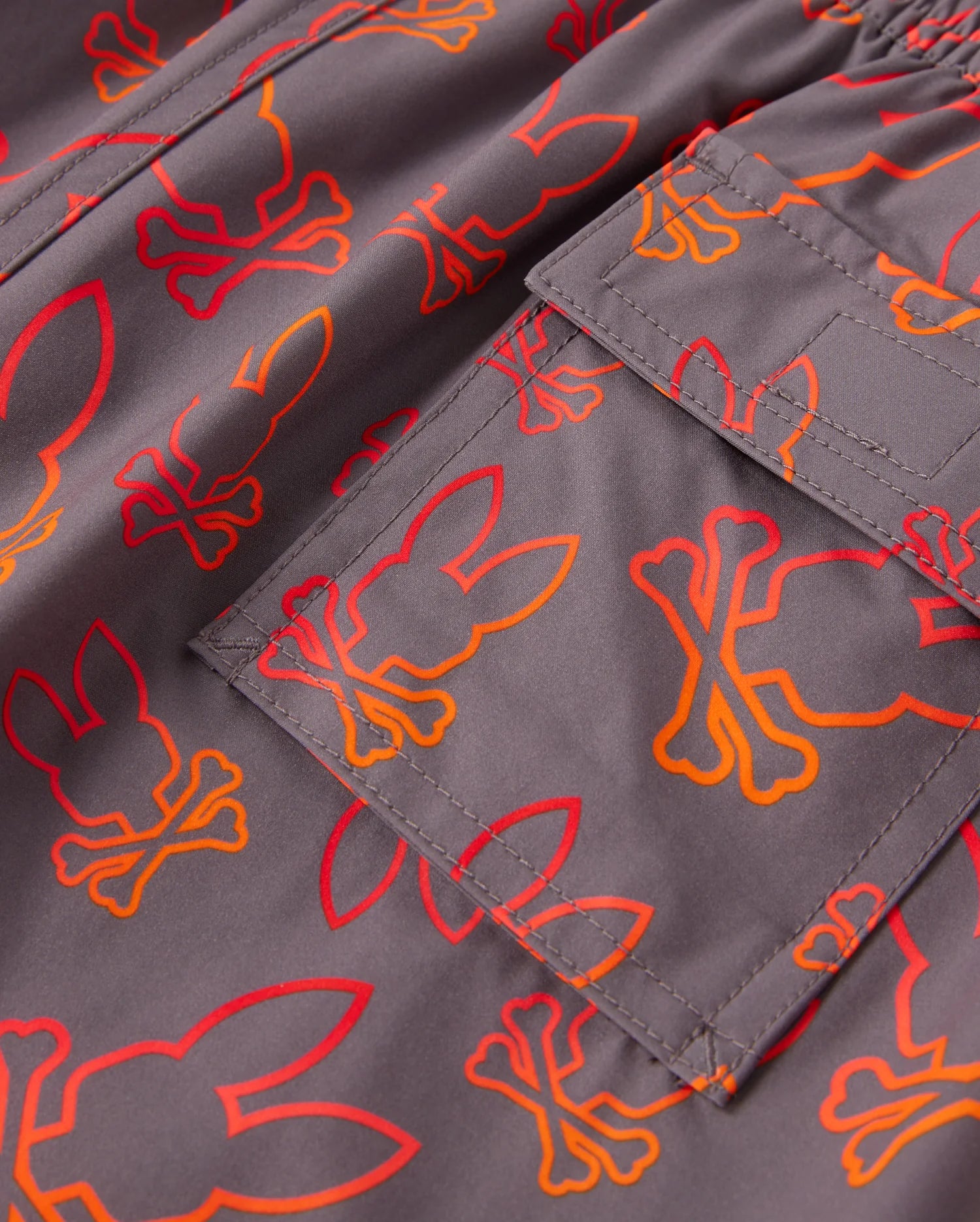 Close-up of a quick-dry fabric with an all-over bunny pattern in orange and red skull-bone designs. The image also highlights a pocket sewn onto the kid's swim trunks, featuring visible stitching and part of the same vibrant pattern. These are the KIDS SHELDON ALL OVER PRINT SWIM TRUNK - B0W588C200 by Psycho Bunny.