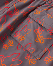 Close-up of a quick-dry fabric with an all-over bunny pattern in orange and red skull-bone designs. The image also highlights a pocket sewn onto the kid's swim trunks, featuring visible stitching and part of the same vibrant pattern. These are the KIDS SHELDON ALL OVER PRINT SWIM TRUNK - B0W588C200 by Psycho Bunny.
