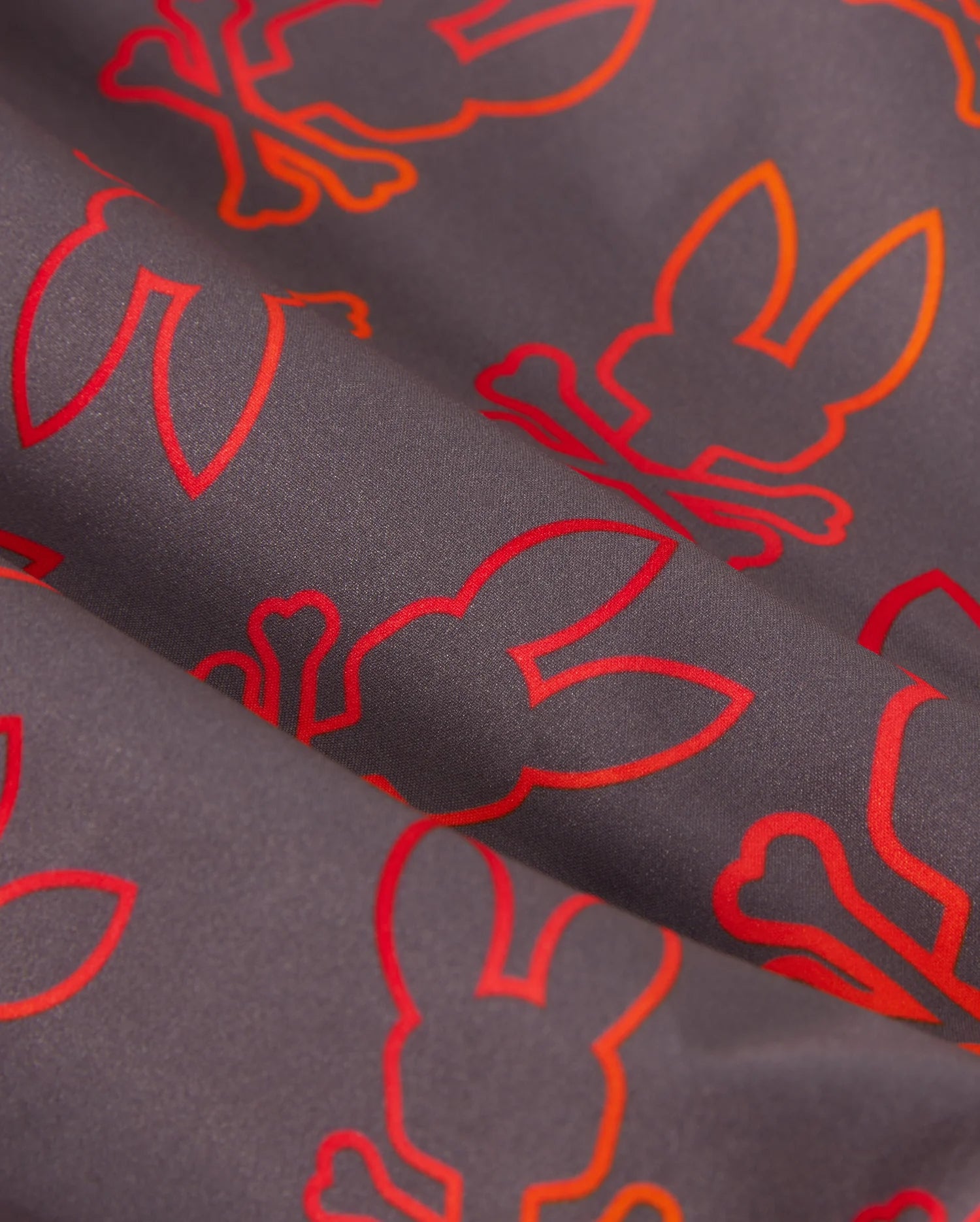 A close-up of a quick-dry fabric with a dark background featuring an orange and red repeating pattern of bunny silhouettes and crossed bones. The bunnies are outlined in bright colors, creating a vibrant contrast with the dark fabric, perfect for kid's swim trunks — the KIDS SHELDON ALL OVER PRINT SWIM TRUNK - B0W588C200 by Psycho Bunny.