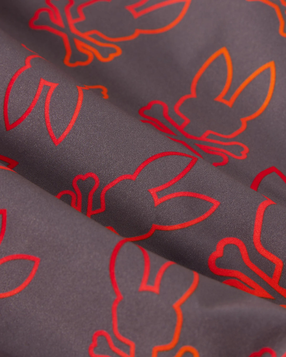 A close-up of a quick-dry fabric with a dark background featuring an orange and red repeating pattern of bunny silhouettes and crossed bones. The bunnies are outlined in bright colors, creating a vibrant contrast with the dark fabric, perfect for kid's swim trunks — the KIDS SHELDON ALL OVER PRINT SWIM TRUNK - B0W588C200 by Psycho Bunny.