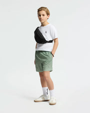 A young boy in green Psycho Bunny KIDS BENDIGO PRINTED SWIM TRUNK - B0W619F200 stands against a white background, wearing a white t-shirt, a black waterproof waist bag across his chest, and white sneakers with tall socks.