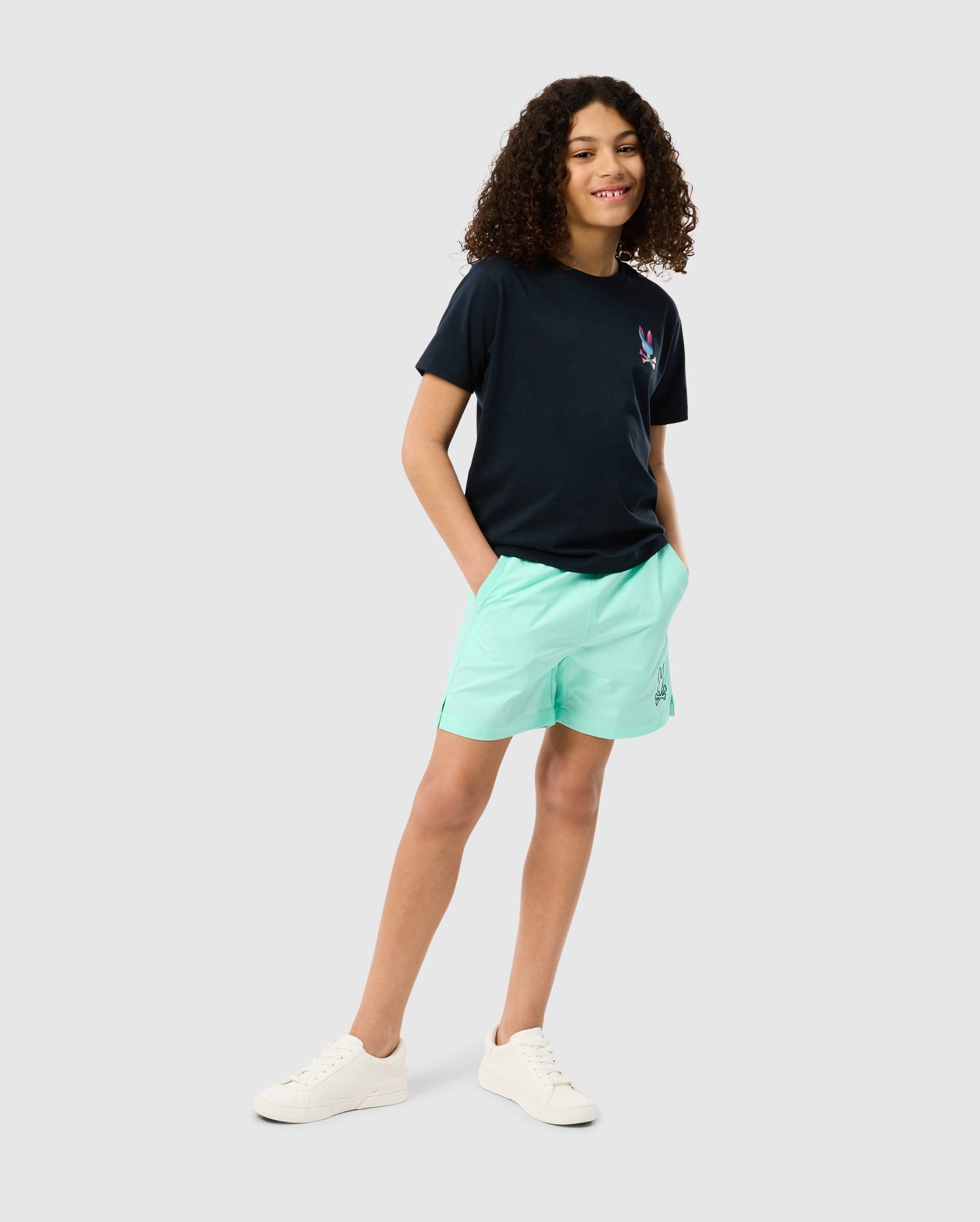 A young person with curly hair wearing a dark blue t-shirt and light green KIDS PARKER HYDROCHROMIC SWIM TRUNK - B0W646C200 by Psycho Bunny stands confidently with hands in pockets. They are also wearing white sneakers and smiling at the camera. The background is plain white.