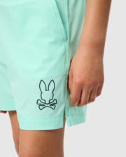Close-up of a person wearing mint green, KIDS PARKER HYDROCHROMIC SWIM TRUNK - B0W646C200 by Psycho Bunny with a black logo of a bunny head above crossed bones on the left leg. The person's right arm is visible, resting alongside their body. The background is plain white.