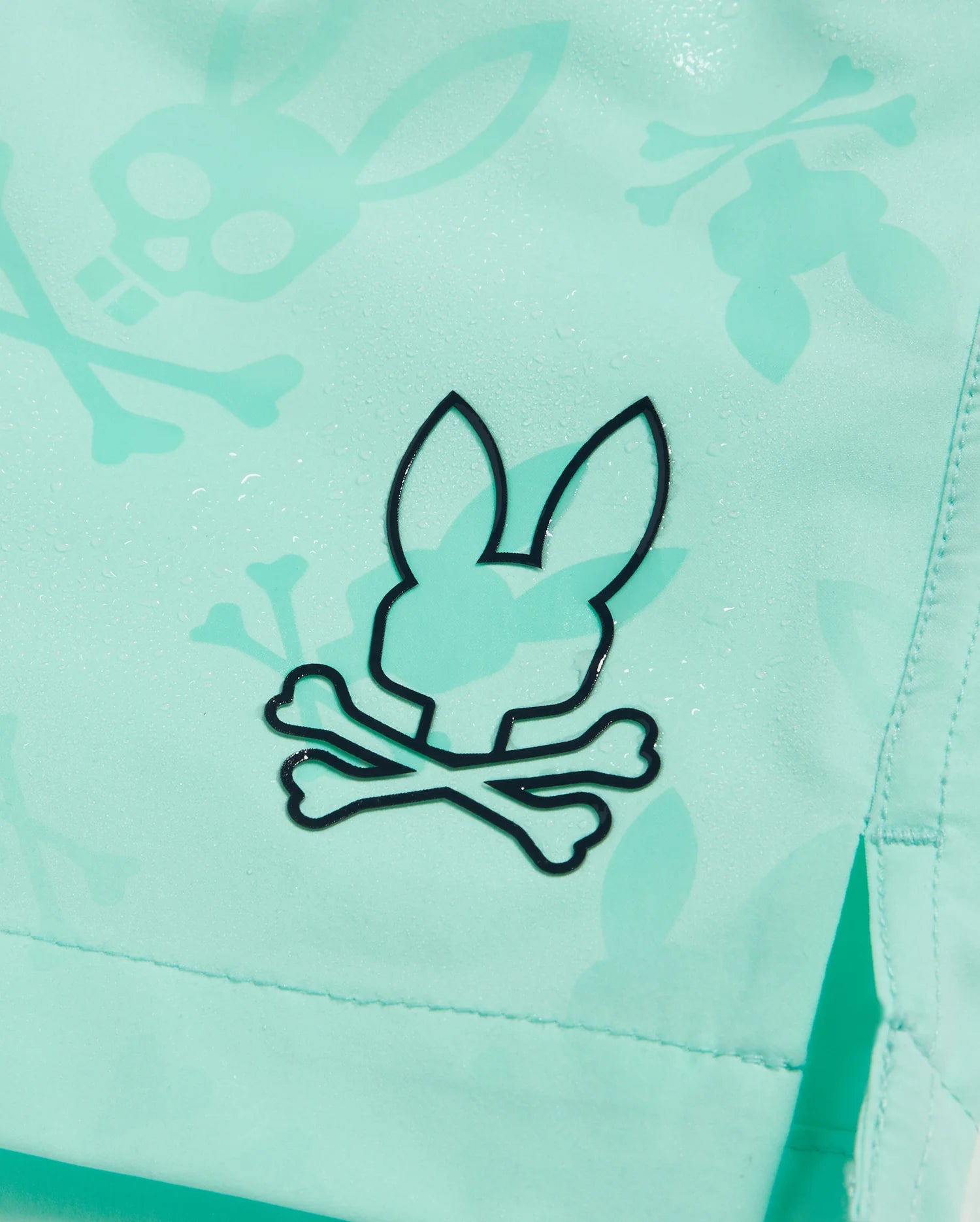 Close-up of a turquoise fabric featuring a black design of a bunny head with crossbones underneath. The Psycho Bunny KIDS PARKER HYDROCHROMIC SWIM TRUNK - B0W646C200 also has a pattern with larger, more translucent bunny and crossbones designs. Water droplets are visible on the fabric.