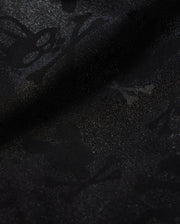 A detailed close-up of a Psycho Bunny KIDS PARKER HYDROCHROMIC SWIM TRUNK - B0W646C200 fabric displaying a subtle textured pattern with skull and crossbones and other abstract shapes. The slightly shiny, quick-dry material adds depth and dimension to the dark hydrochromatic pattern.