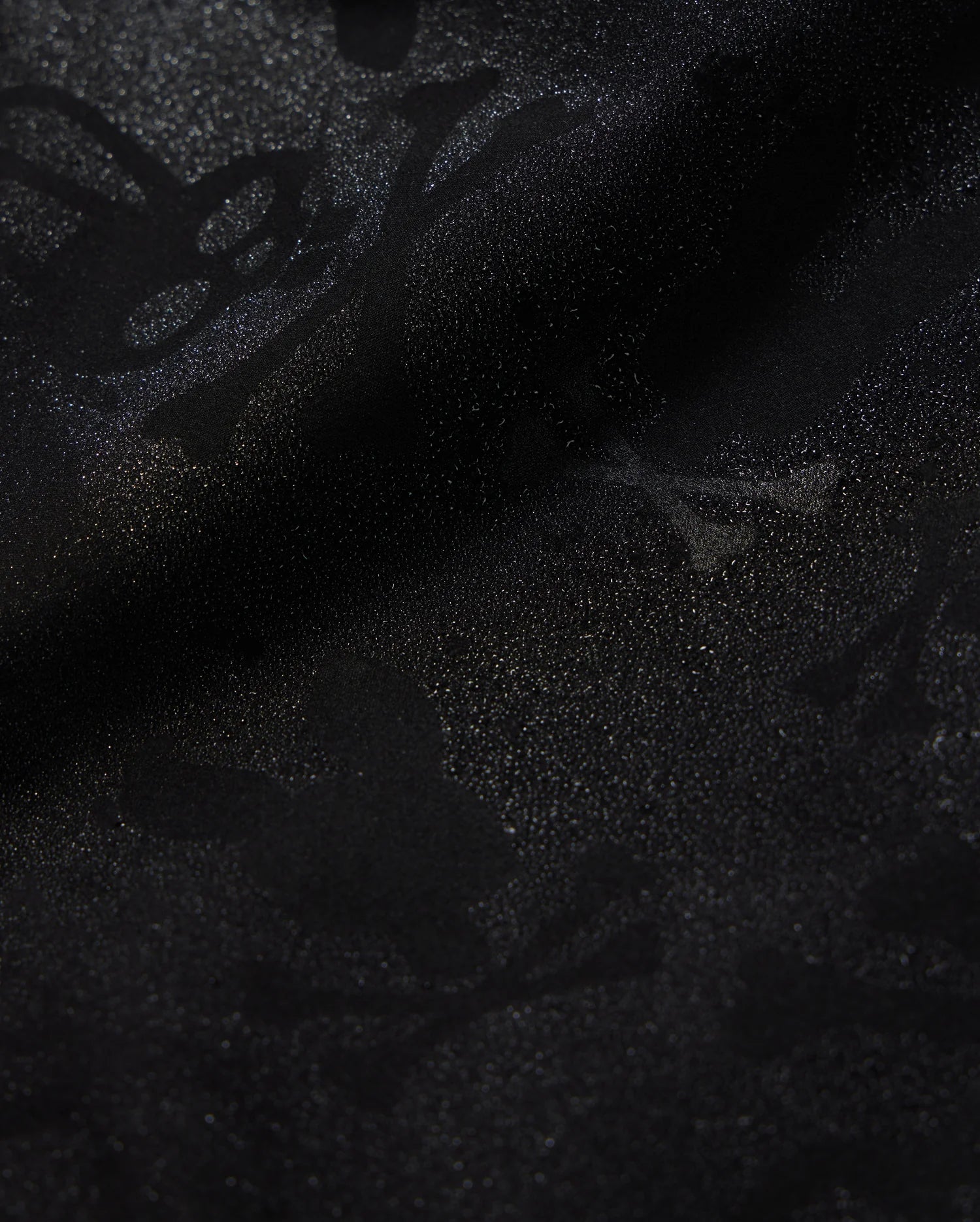 A detailed close-up of a Psycho Bunny KIDS PARKER HYDROCHROMIC SWIM TRUNK - B0W646C200 fabric displaying a subtle textured pattern with skull and crossbones and other abstract shapes. The slightly shiny, quick-dry material adds depth and dimension to the dark hydrochromatic pattern.