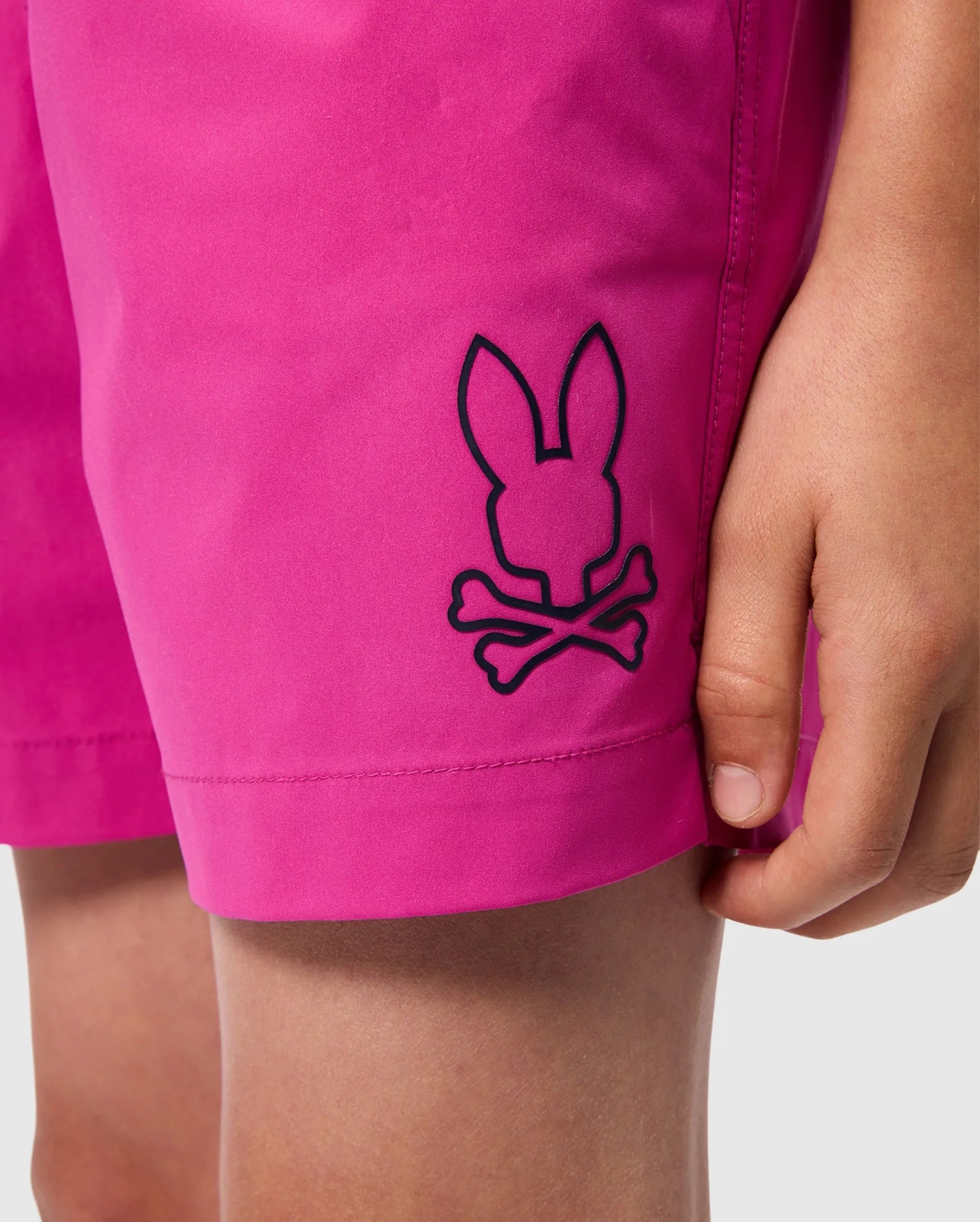 Close-up of a person wearing the KIDS PARKER HYDROCHROMIC SWIM TRUNK - B0W646C200 by Psycho Bunny, showcasing its bright pink, quick-drying fabric with a black bunny pattern near the hem. The person’s hand is visible resting against the shorts.