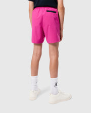 A person standing with their back to the camera, wearing bright pink Psycho Bunny KIDS PARKER HYDROCHROMIC SWIM TRUNKS (B0W646C200), a black shirt, white socks, and white sneakers. The shorts feature a black pocket on the right side. The background is plain and gray.