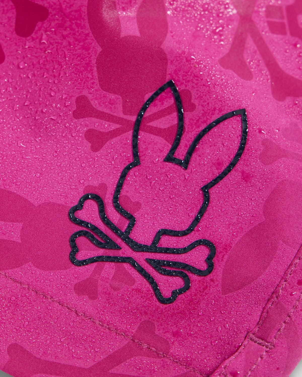 Close-up of a pink fabric with water droplets on its surface, featuring a black outlined design of a bunny's head with crossbones below it. The background includes a repeated bunny pattern in a slightly darker shade of pink, perfect for the quick-drying KIDS PARKER HYDROCHROMIC SWIM TRUNK - B0W646C200 by Psycho Bunny.