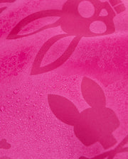 A close-up of the textured pink fabric of Psycho Bunny's KIDS PARKER HYDROCHROMIC SWIM TRUNK - B0W646C200, featuring a bunny pattern with long ears and skull-like faces. Some areas have visible water droplets, emphasizing the hydrochromic swim trunks' quick-drying properties.