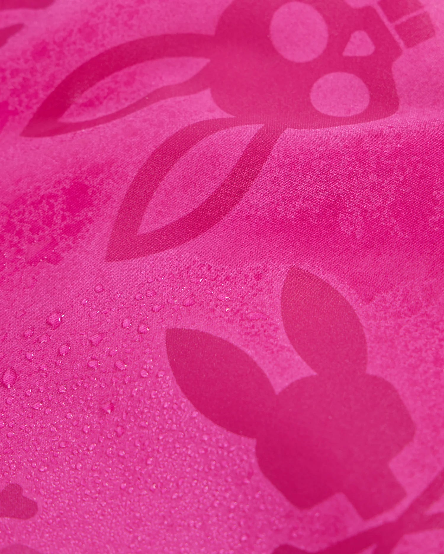 A close-up of the textured pink fabric of Psycho Bunny's KIDS PARKER HYDROCHROMIC SWIM TRUNK - B0W646C200, featuring a bunny pattern with long ears and skull-like faces. Some areas have visible water droplets, emphasizing the hydrochromic swim trunks' quick-drying properties.