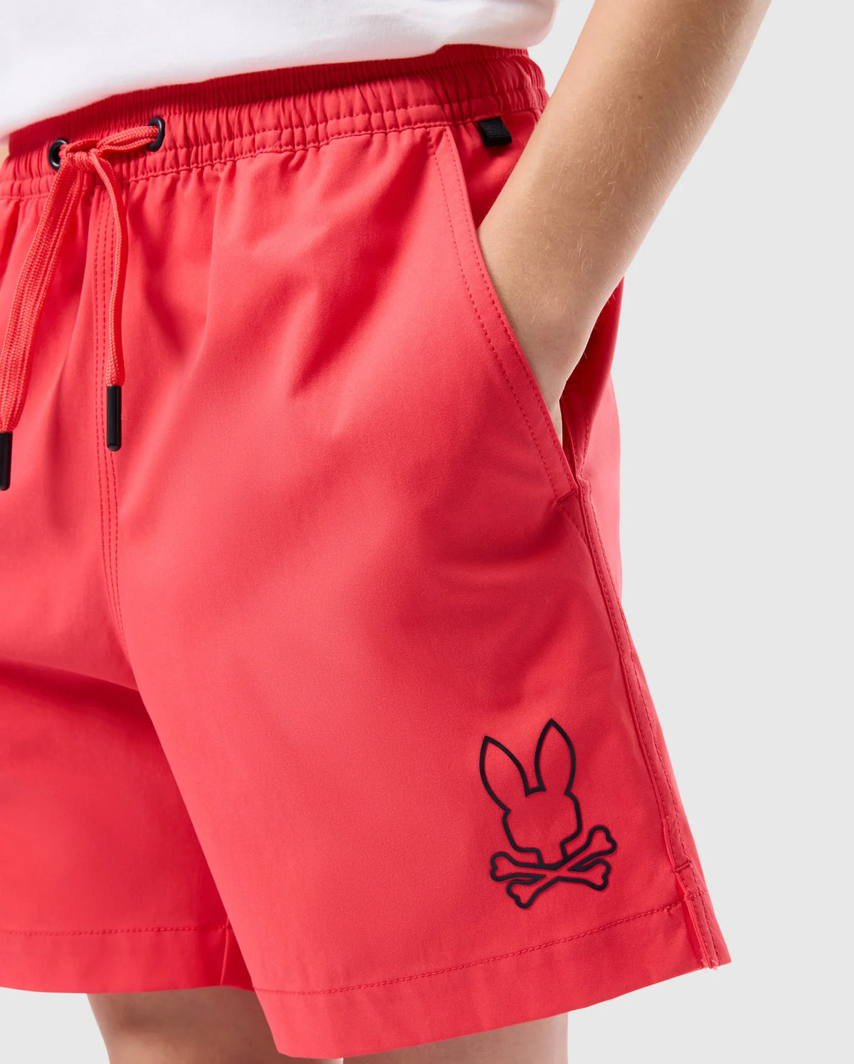 A person wearing bright red, quick-dry material shorts with their hand in the pocket is shown. The shorts feature an embroidered black logo of a skull and crossbones, where the skull is stylized as a rabbit's head. The white shirt tucked into the shorts is partially visible. These are the Psycho Bunny KIDS PARKER HYDROCHROMIC SWIM TRUNK - B0W646C200.