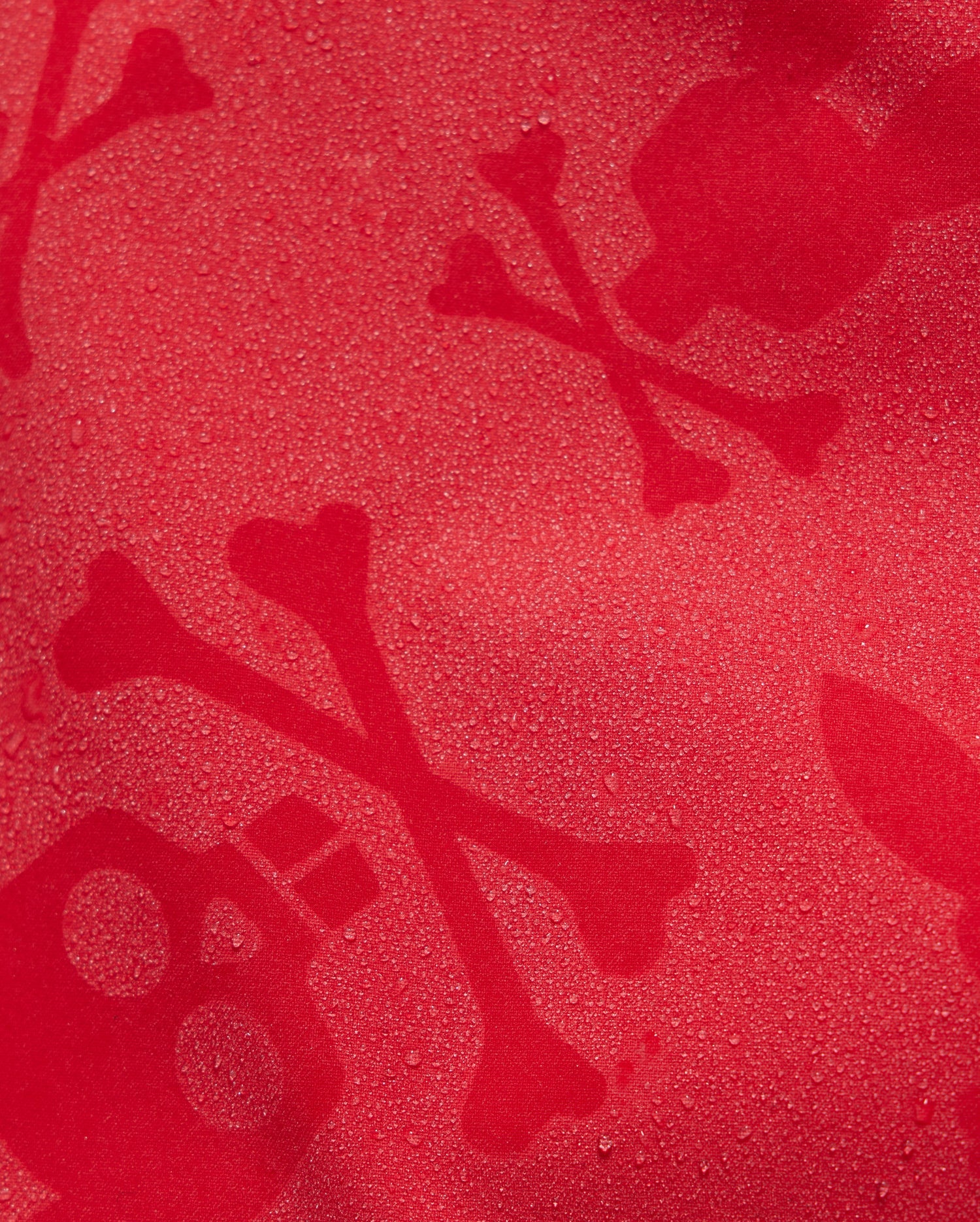 A close-up of a red KIDS PARKER HYDROCHROMIC SWIM TRUNK - B0W646C200 by Psycho Bunny with moisture droplets forming a pattern that includes skulls and crossbones. The skulls have two circular eyes and a rectangular nose, while the crossbones intersect with two bones each.