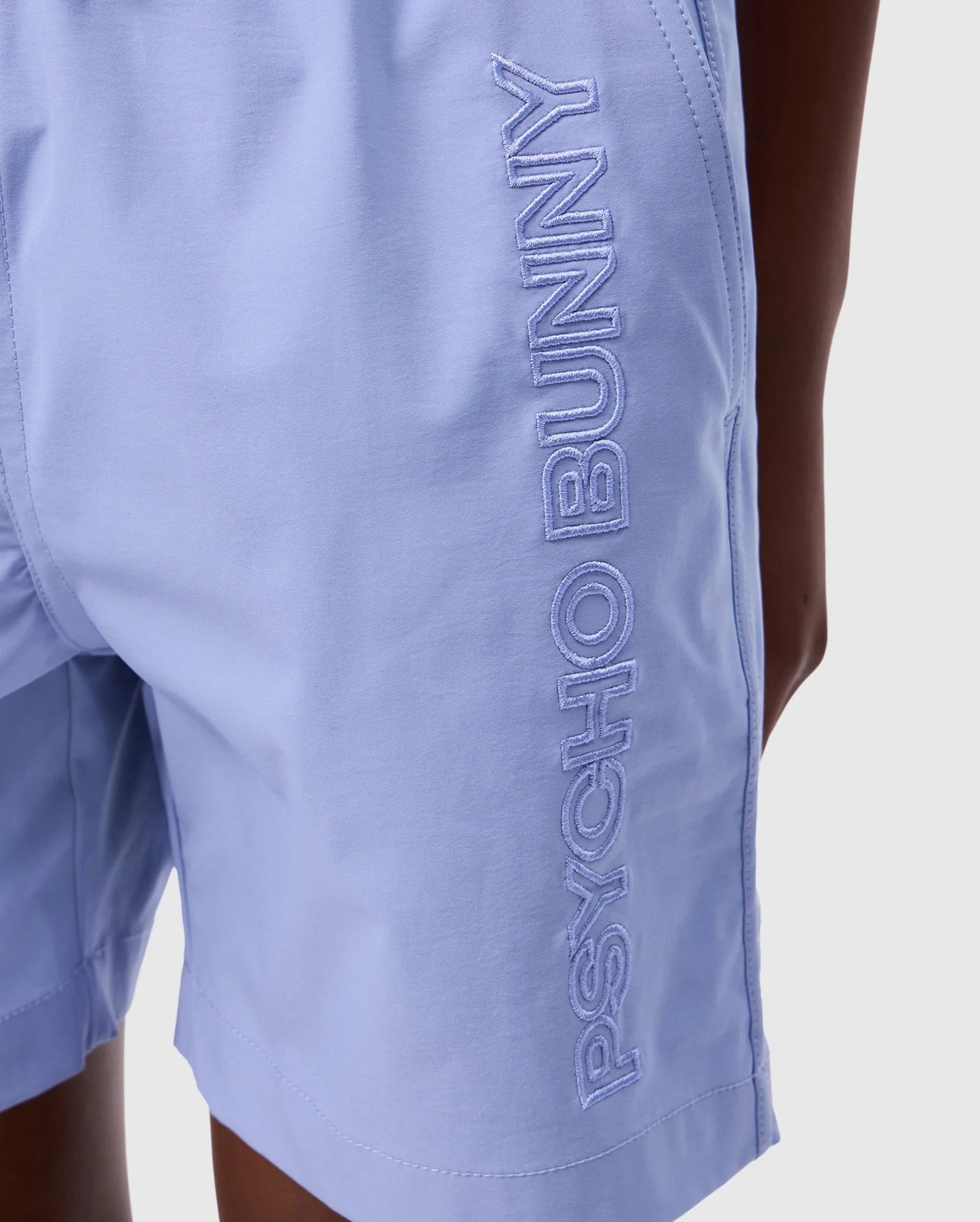 Close-up of a person wearing light blue KIDS MELVIN SWIM TRUNK - B0W665C200 with the words "PSYCHO BUNNY" embroidered in large white letters on the side. The stretch mesh fabric ensures comfort, and the person's right hand rests near their hip. The background is plain white.