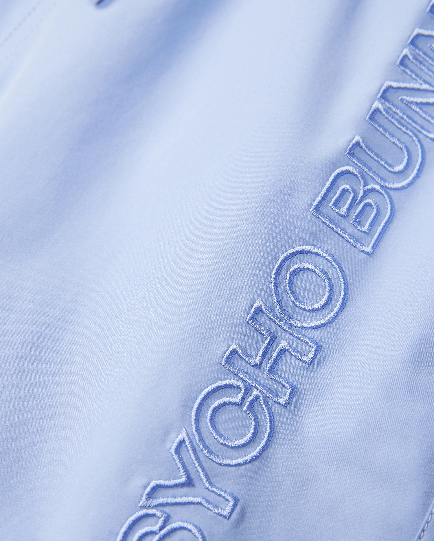 The product is a light blue KIDS MELVIN SWIM TRUNK (B0W665C200) by Psycho Bunny, featuring "PSYCHO BUN" partially embroidered in blue thread. This quick-dry fabric ensures comfort and style by the water.