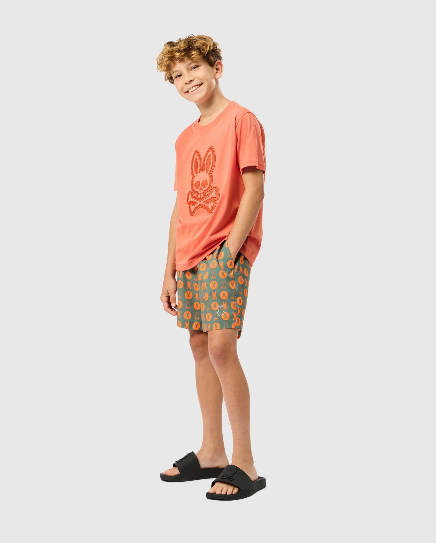A young boy with curly hair stands smiling, wearing an orange t-shirt featuring a bunny graphic and the Psycho Bunny KIDS ARNOLD PRINT SWIM TRUNK (B0W820D200). With his hands in his pockets, he poses against a plain light gray background, showcasing black sandals for an effortless style.