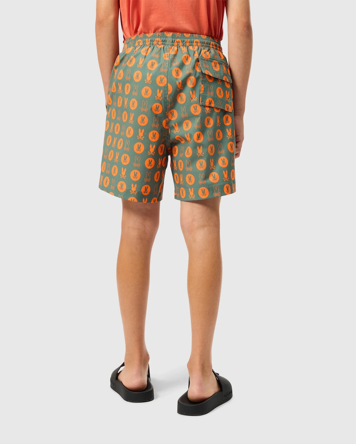 A person is seen from behind, dressed in the vibrant KIDS ARNOLD PRINT SWIM TRUNK by Psycho Bunny, which showcases lively orange and green patterns. These trunks feature a quick-dry fabric and a stretch mesh lining. They are also wearing an orange shirt paired with black sandals.