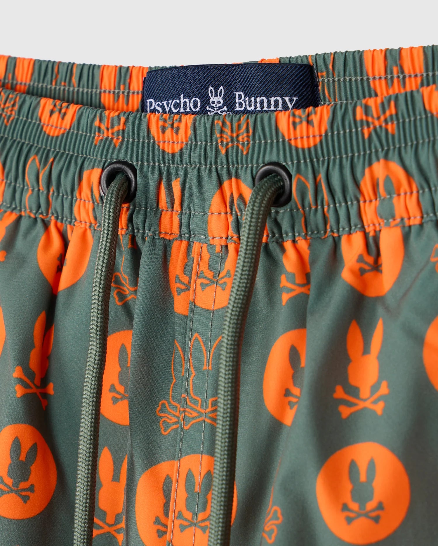 Close-up of the KIDS ARNOLD PRINT SWIM TRUNK - B0W820D200 featuring green swim trunks with orange patterns, including a bunny and crossed bones. The quick-dry material ensures comfort, complemented by a waistband adorned with a black Psycho Bunny label. These trunks are designed with stretch mesh lining and green drawstrings for the perfect fit.