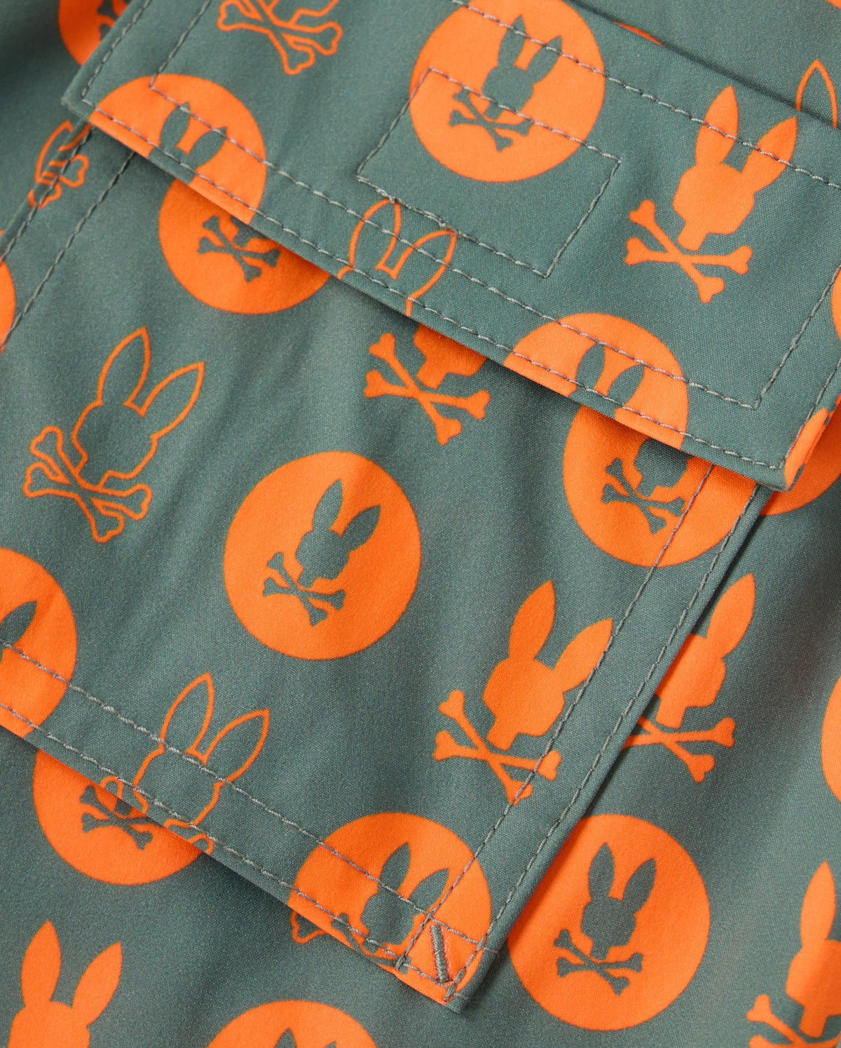 A close-up view of the Psycho Bunny KIDS ARNOLD PRINT SWIM TRUNK (B0W820D200) showcases its green fabric with an orange pattern of bunny heads and crossbones inside circles. The swim trunk features a center pocket with visible stitching and is crafted from quick-dry material for ultimate comfort and convenience.