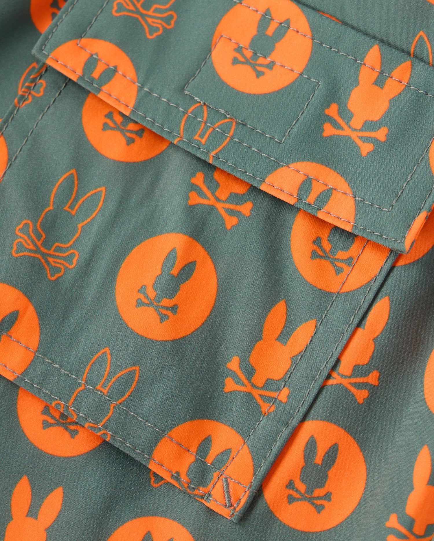 A close-up view of the Psycho Bunny KIDS ARNOLD PRINT SWIM TRUNK (B0W820D200) showcases its green fabric with an orange pattern of bunny heads and crossbones inside circles. The swim trunk features a center pocket with visible stitching and is crafted from quick-dry material for ultimate comfort and convenience.
