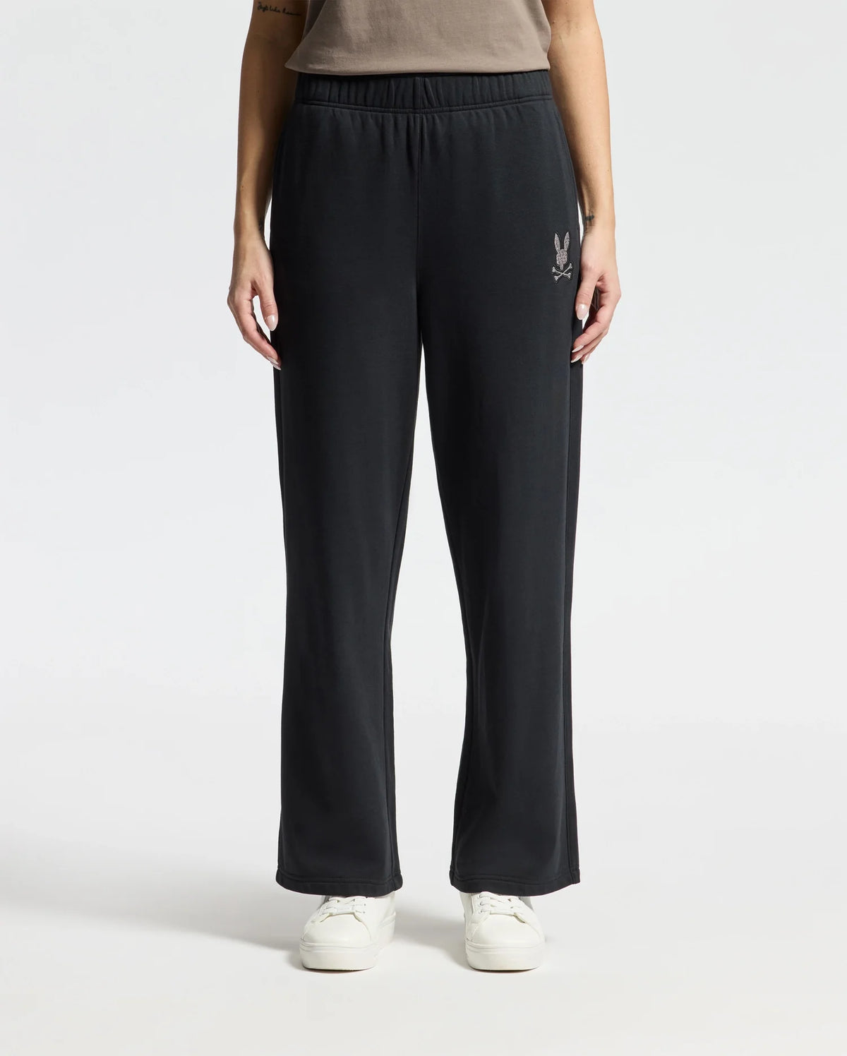 A person sporting the stylish Psycho Bunny WOMENS ALEXANDER SWEATPANT - B2P157E200, featuring a subtle logo on the left thigh. The black sweatpants are complemented by white sneakers against a simple, neutral backdrop.
