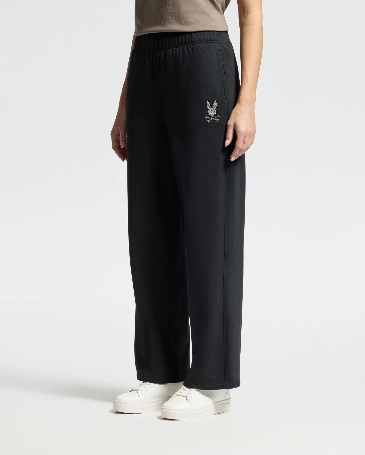 A person wearing the stylish WOMENS ALEXANDER SWEATPANT - B2P157E200 by Psycho Bunny, featuring an emblem on the left side, and white sneakers stands against a plain background. The person is partially visible from the waist down.