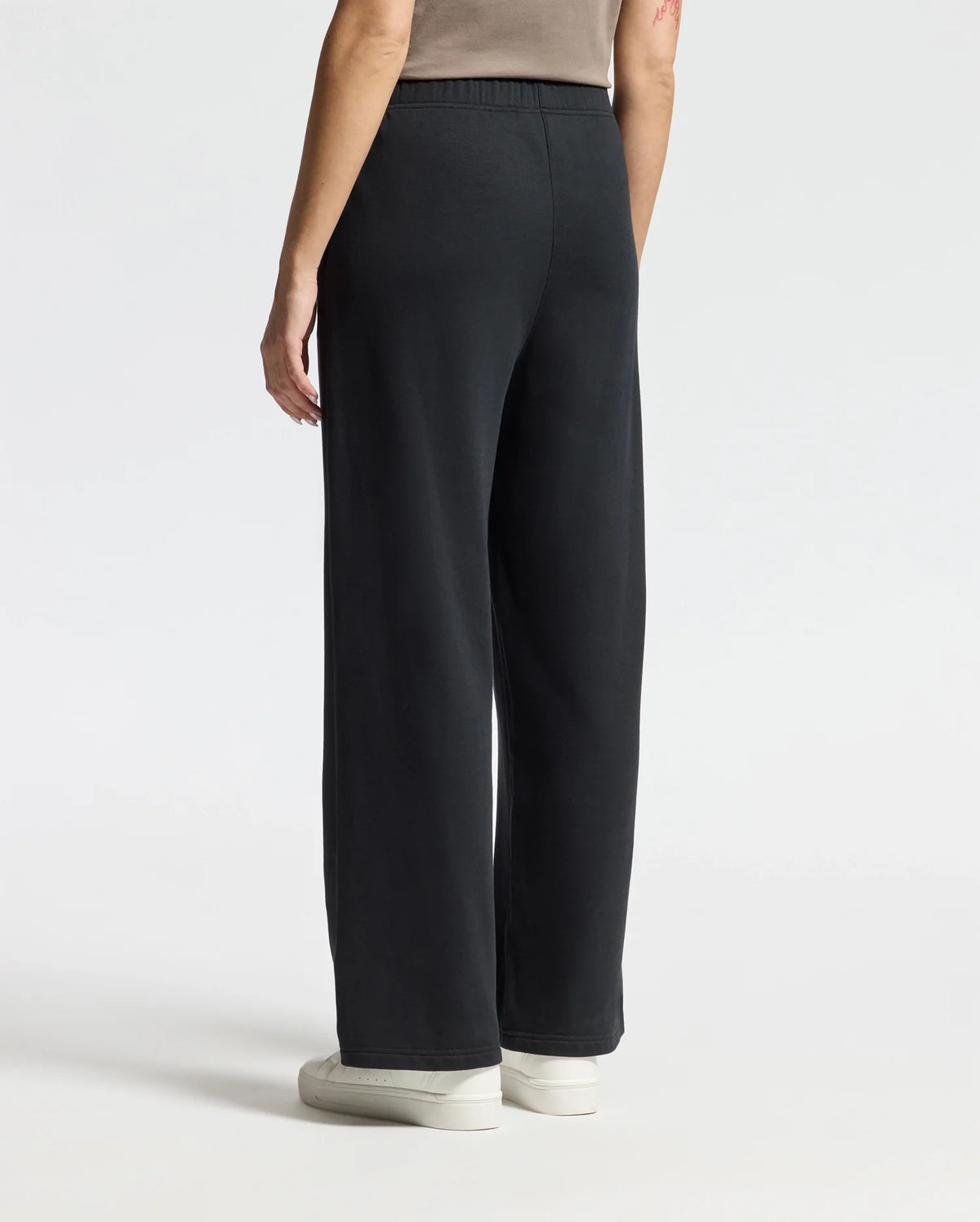 A person wearing stylish Psycho Bunny WOMENS ALEXANDER SWEATPANT - B2P157E200 and white sneakers is standing with their back facing the camera against a plain, light-colored background.