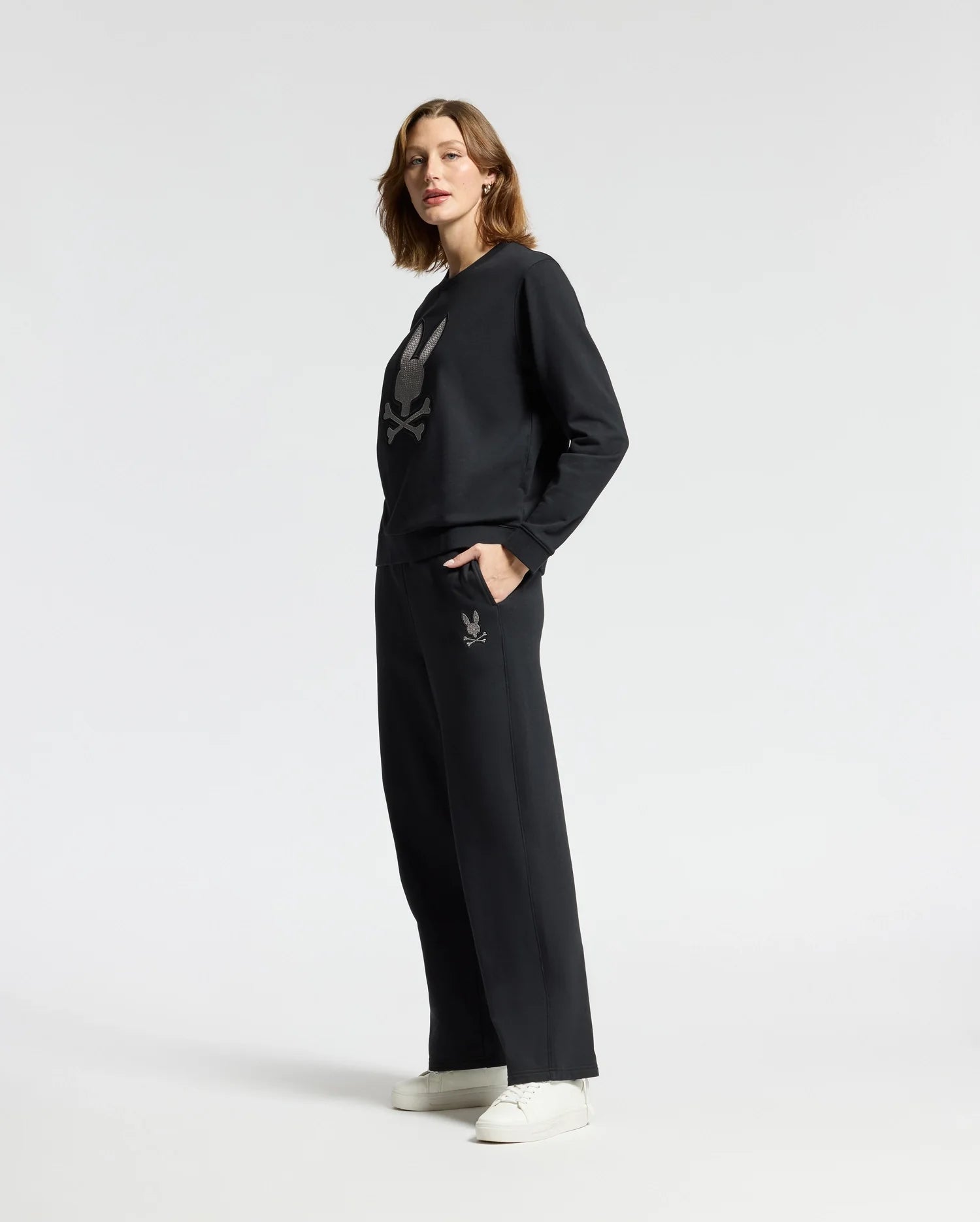 A person models the Psycho Bunny WOMENS ALEXANDER SWEATPANT - B2P157E200 in black featuring a graphic design on the front, set against a white backdrop. They have their hands comfortably in the pockets of these stylish cotton blend sweatpants and are paired with white sneakers.