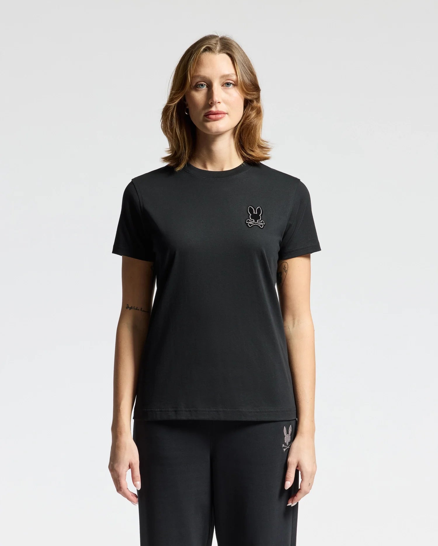 A woman wearing the Psycho Bunny WOMENS ALEXANDER GRAPHIC TEE (B2U164E200), crafted from soft Pima cotton, and black pants stands against a plain white background. The tee is adorned with a small rabbit logo featuring delicate lurex embroidery on the left chest.