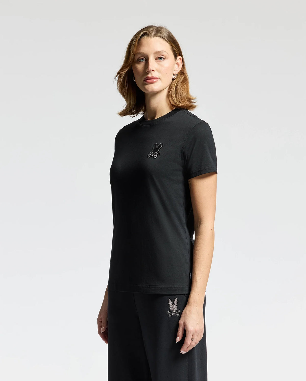 A woman with shoulder-length brown hair confidently gazes at the camera while wearing a stylish black Pima Cotton t-shirt, identified as the Psycho Bunny WOMENS ALEXANDER GRAPHIC TEE - B2U164E200, along with matching pants. The outfit is adorned with a delicate rabbit graphic on both the chest and leg. She stands against a light gray background.
