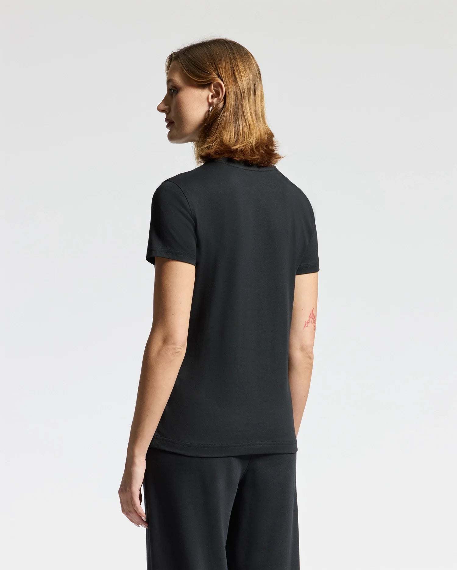 A person with shoulder-length hair is standing in a neutral pose, facing left. They are wearing a black WOMENS ALEXANDER GRAPHIC TEE by Psycho Bunny and pants. The background is plain white.