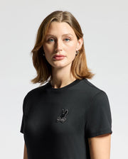 A person with shoulder-length brown hair is wearing a stylish Psycho Bunny Women's Alexander Graphic Tee (B2U164E200) in Pima Cotton, featuring a small Lurex Embroidery design on the chest. The background is plain and light-colored.